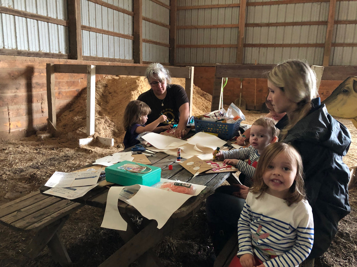 Two adults and four young children doing a paper-and-glue craft at a weathered picnic table inside a pole barn, a painted plywood cow cut-out against the wall