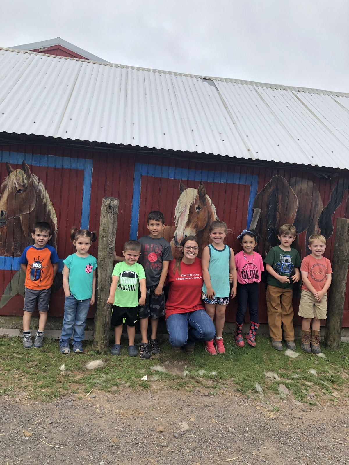 Eight young children lined up along the mural-painted red barn wall with an adult in a farm staff t-shirt kneeling in the middle of the group
