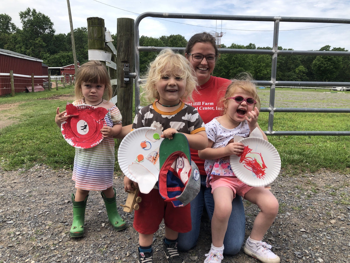 Three small children holding up paper-plate animal crafts they have painted, an adult in a farm staff t-shirt kneeling behind them