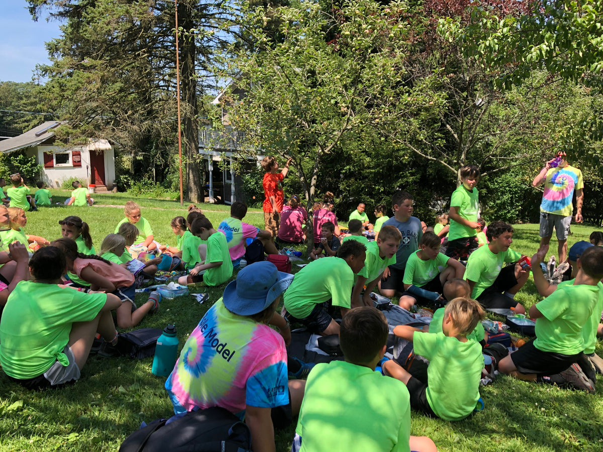 A large group of children in matching green camp t-shirts sitting on the lawn under fruit trees eating packed lunches