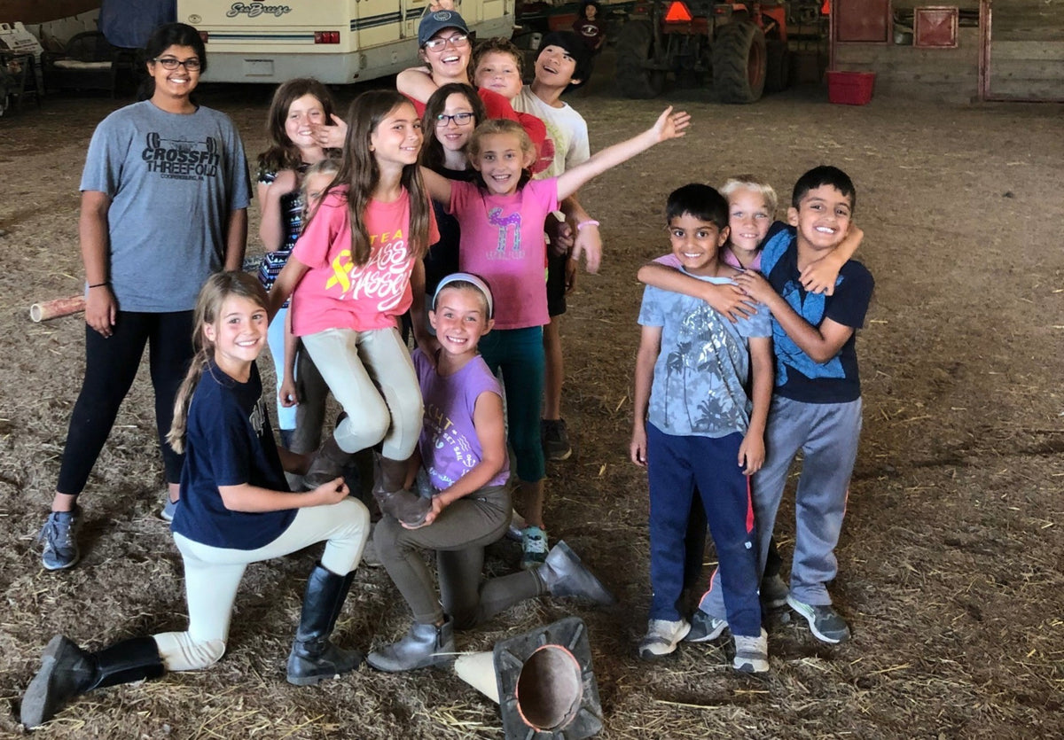 A group of children and teenagers laughing together in the indoor riding arena, several in riding breeches and boots