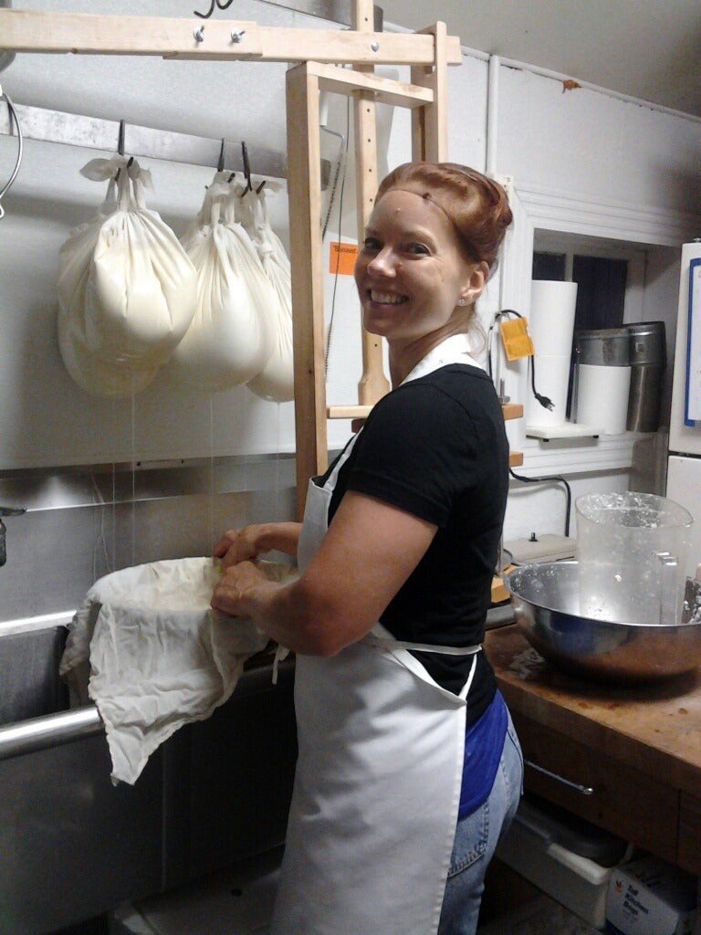 An adult lifting a cheesecloth-lined form over a sink, bags of curd draining behind
