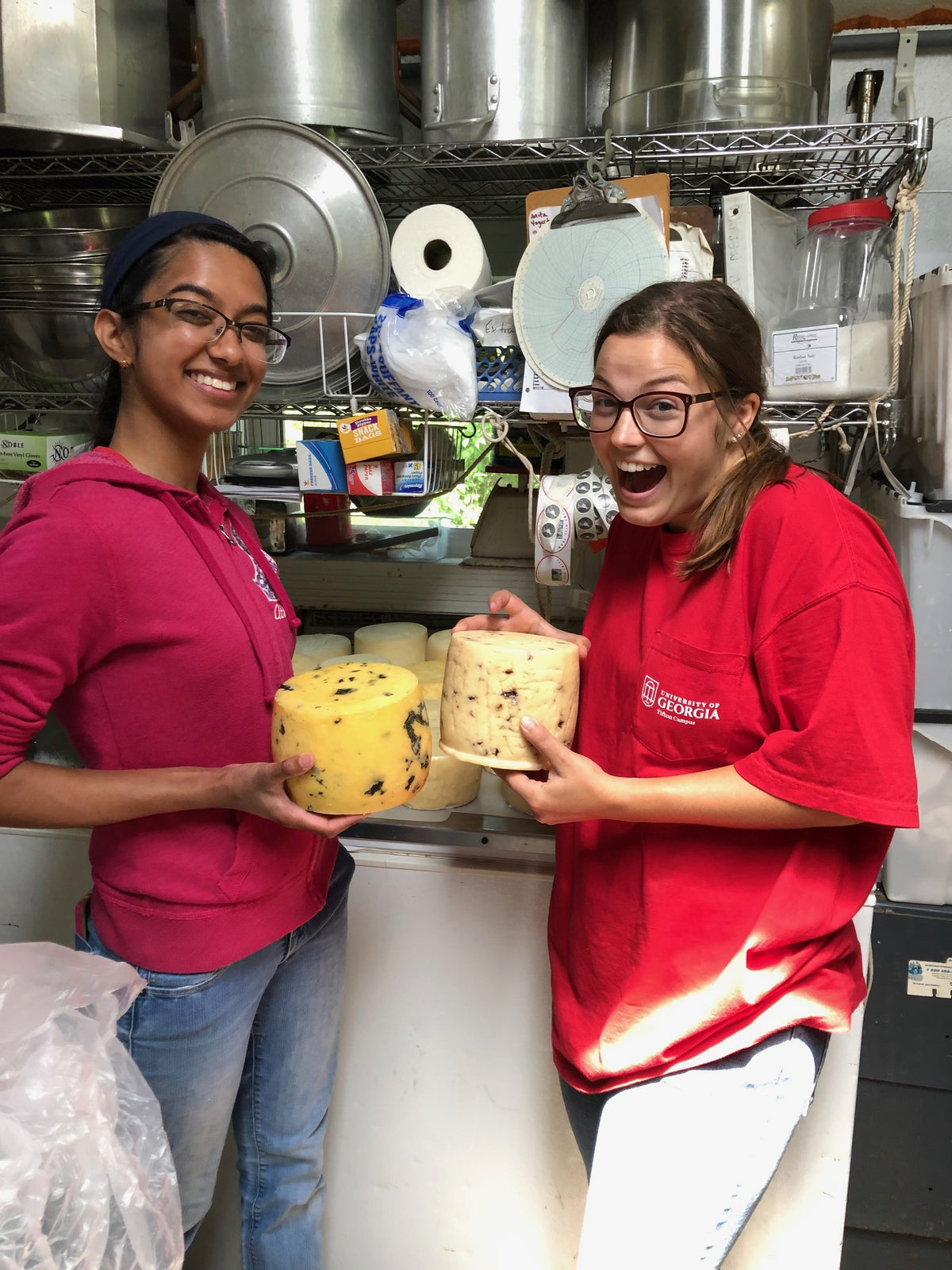 Two people in the cheese room holding up aged wheels studded with dark inclusions, racks behind