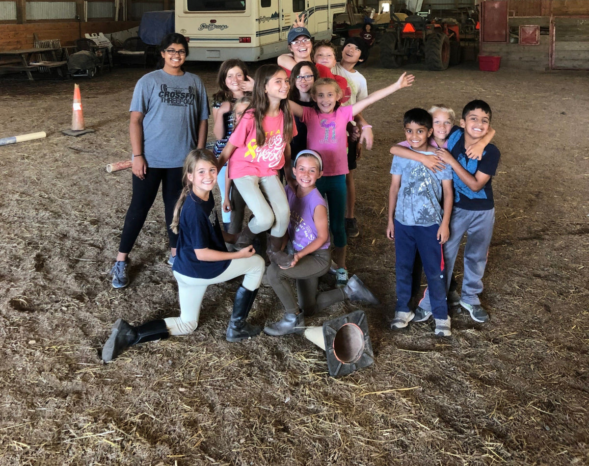 About thirteen children posed with two young leaders in the indoor riding arena, two in breeches and tall boots kneeling in front