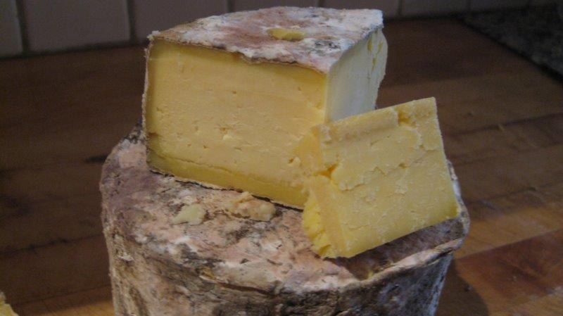 An aged natural-rind cheese wheel on a wooden counter with a wedge cut out, showing dense pale-yellow paste and a mottled gray-white rind