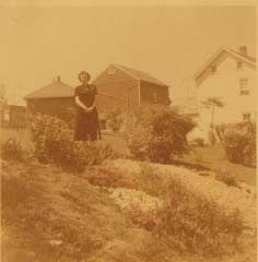 Sepia archival photograph of a woman in a long dark dress in the hillside garden below the farm buildings