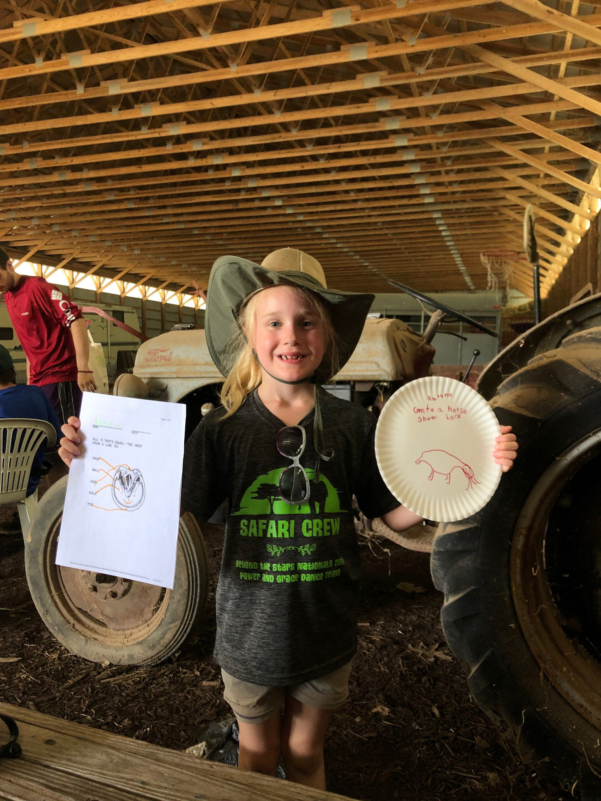 Barn classroom by a tractor: a person in a sun hat holds up a paper-plate pig while another holds a worksheet on the parts of a hoof