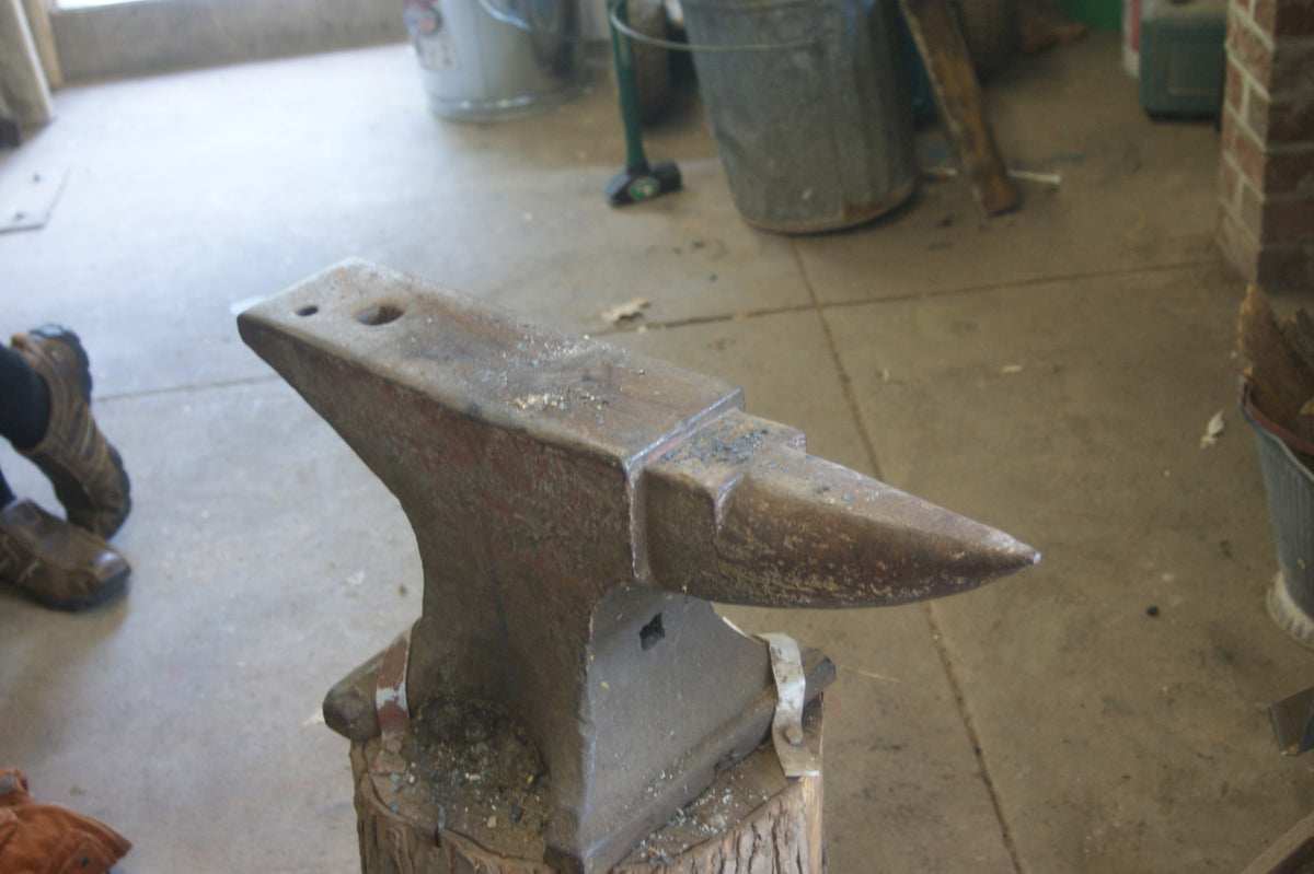A blacksmith's anvil on a wooden stump on a concrete shop floor, scale flakes on its face, galvanized buckets, a sledge and tools behind