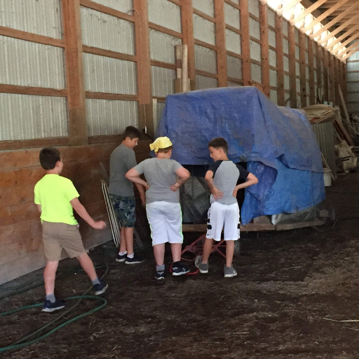 Four boys standing in a pole barn looking at a hay wagon covered with a blue tarp, a garden hose coiled on the dirt floor