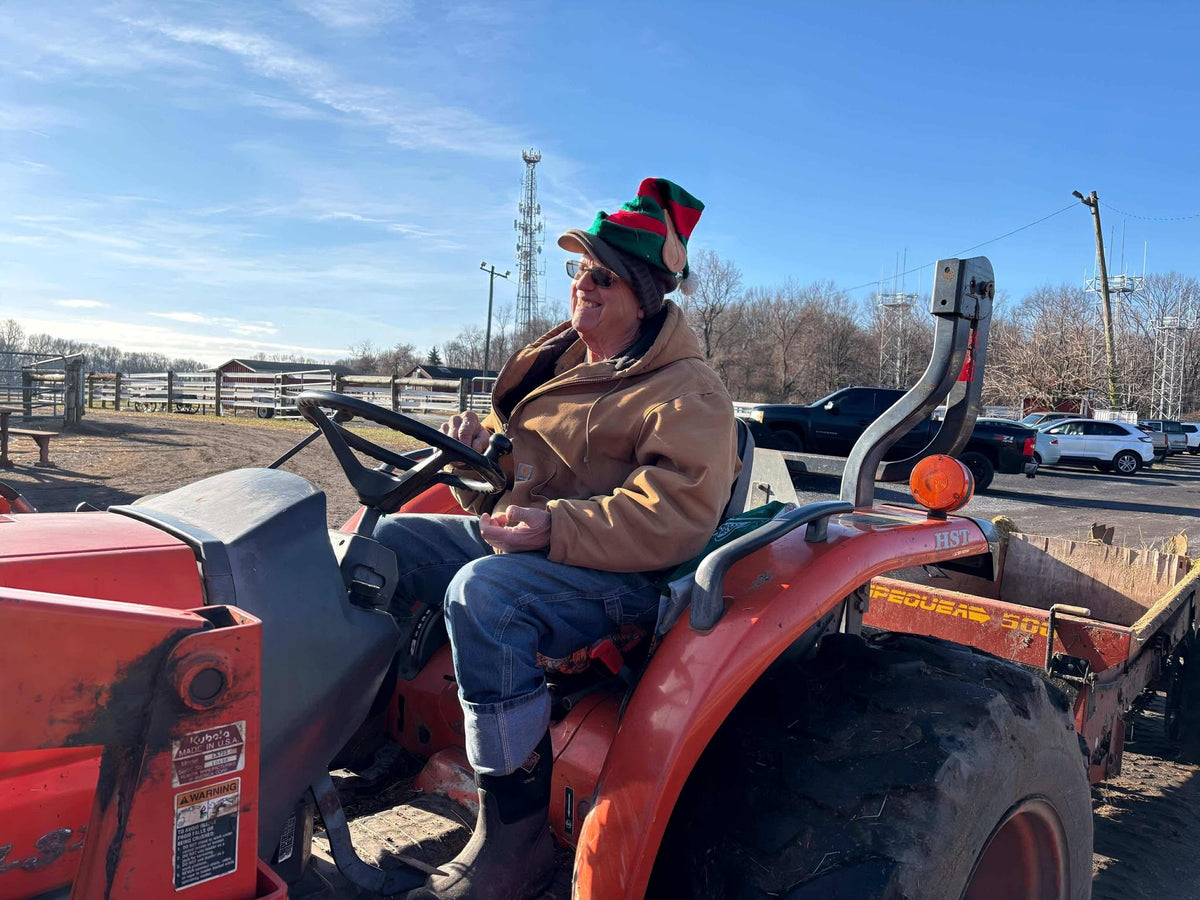 An adult in a Carhartt jacket and red-and-green elf hat at the wheel of an orange Kubota tractor, smiling off to the side