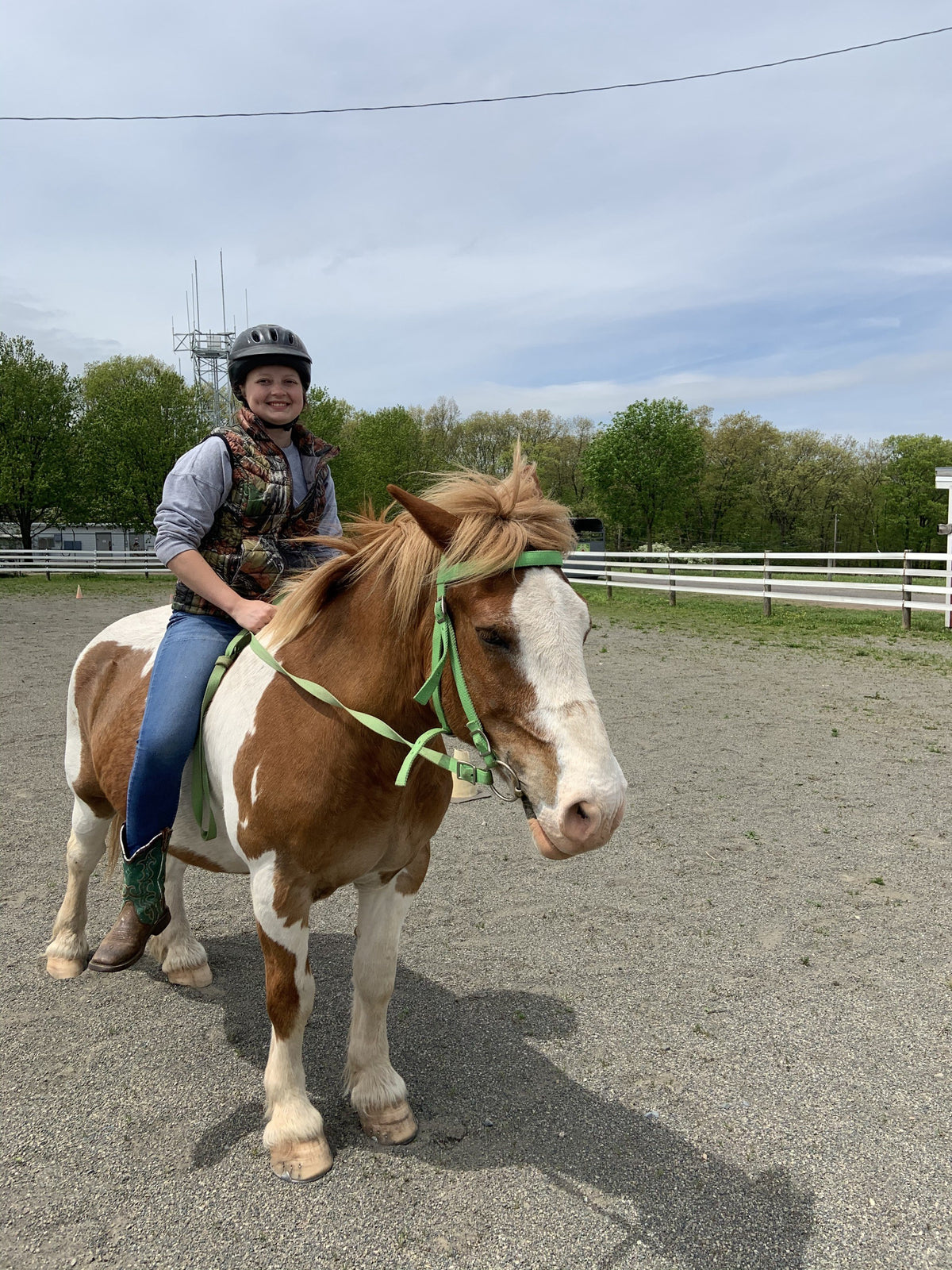 A child in a riding helmet and camo vest sitting bareback on a chestnut-and-white pinto pony in a sand riding ring, spring trees behind