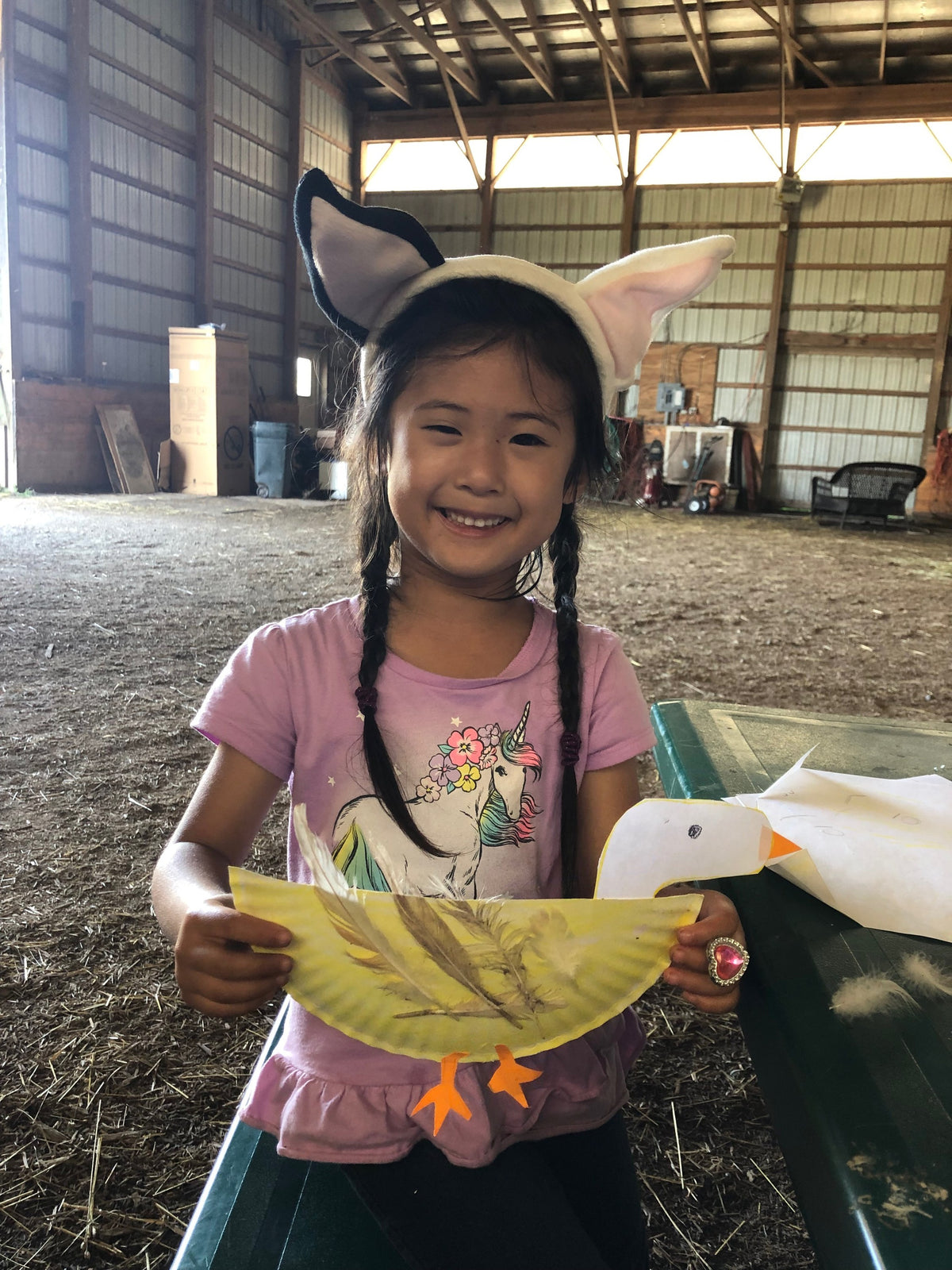 A young child in a felt animal-ear hood grinning in the barn, holding up a paper-plate duck made with real feathers and orange paper feet