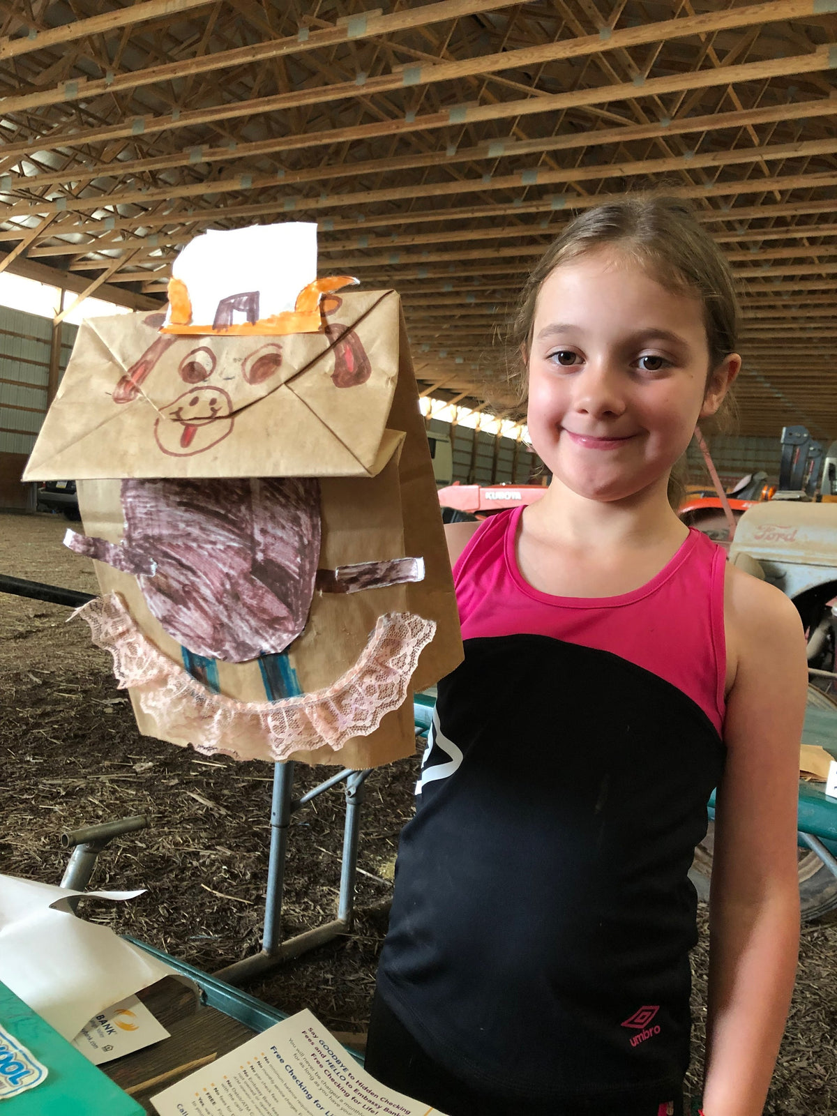 A child in the barn holding up a paper-bag cow puppet decorated in crayon with lace trim, made during a craft activity