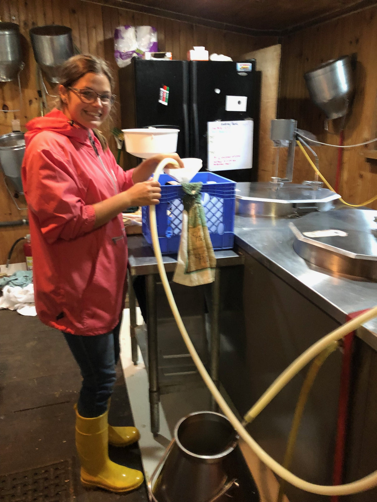 A child in a pink raincoat and yellow rain boots handling milk line tubing in the dairy room, beside stainless milk cans and a bulk tank