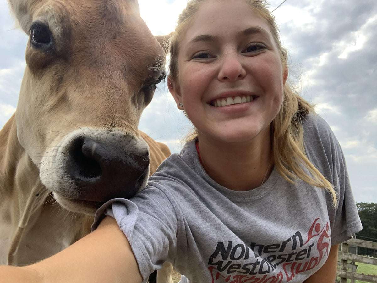 Close selfie of a young woman cheek-to-cheek with a fawn Jersey cow, her arm around its neck, in a pasture under a bright overcast sky