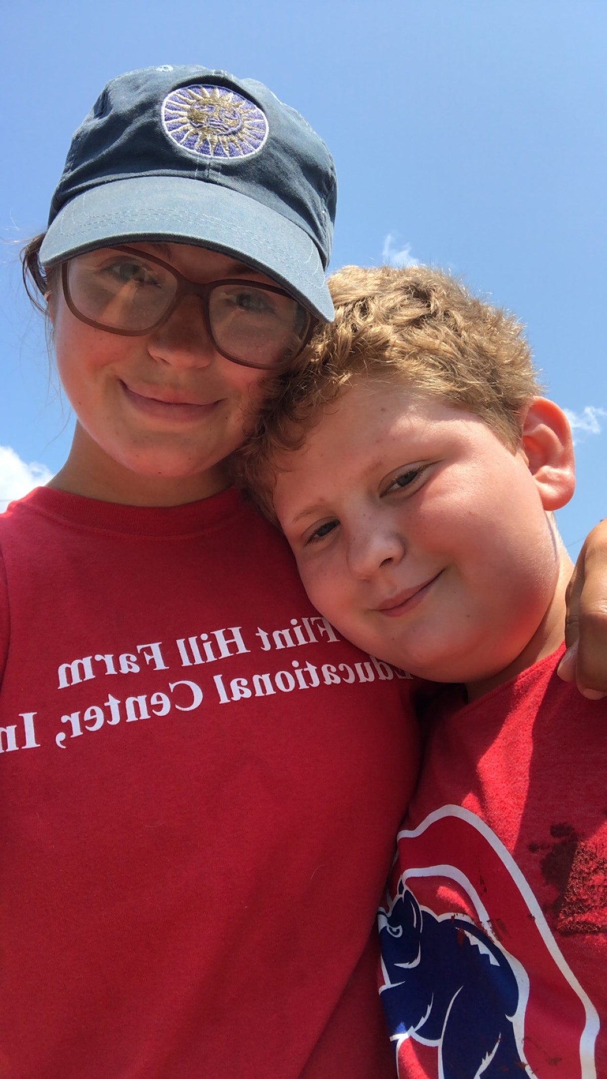 Outdoor selfie of a teenager in a red Flint Hill Farm Educational Center T-shirt and cap, arm around a younger child, against blue sky