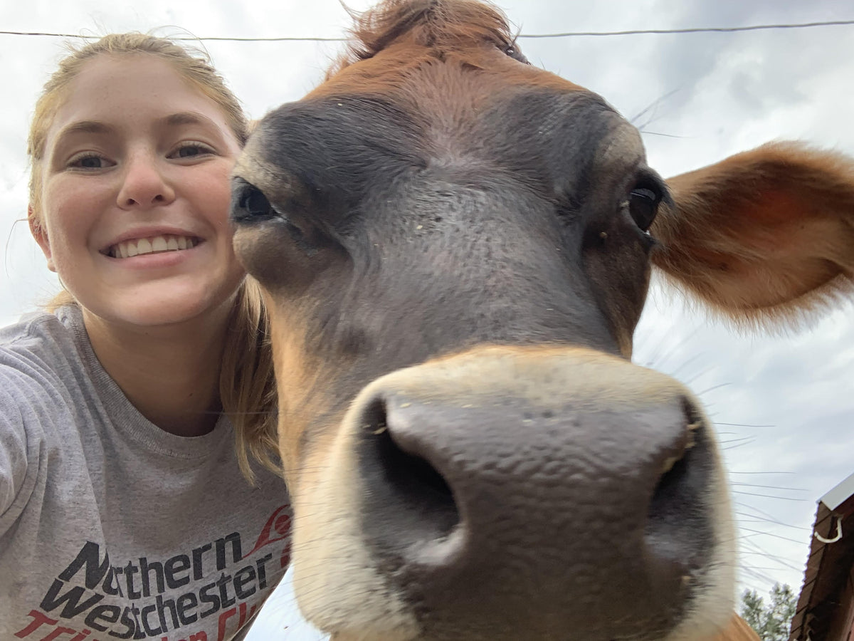 Close-up selfie of a young woman cheek-to-cheek with a Jersey cow, the cow's muzzle filling most of the frame under an overcast sky