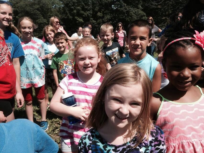 A crowd of school-age children pressed close to the camera outdoors on a sunny field trip, several adults standing behind them