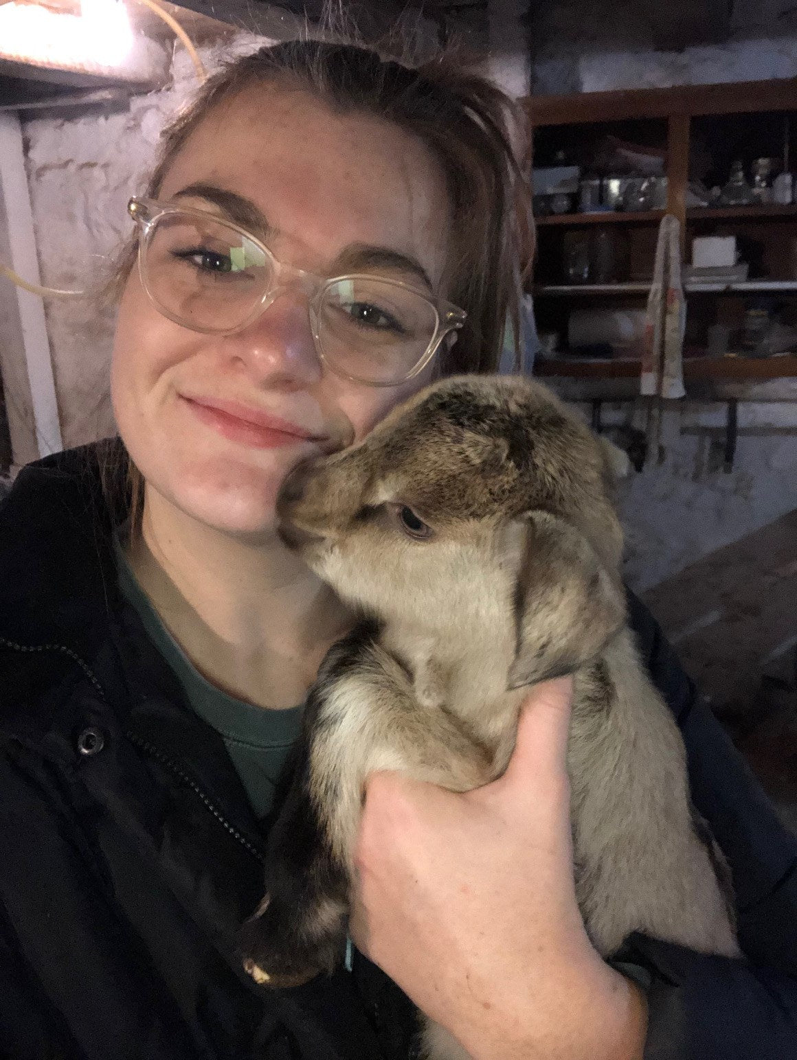 An adult in a dark jacket and glasses holding a young goat kid against her cheek and smiling, in a dim stone-walled barn room