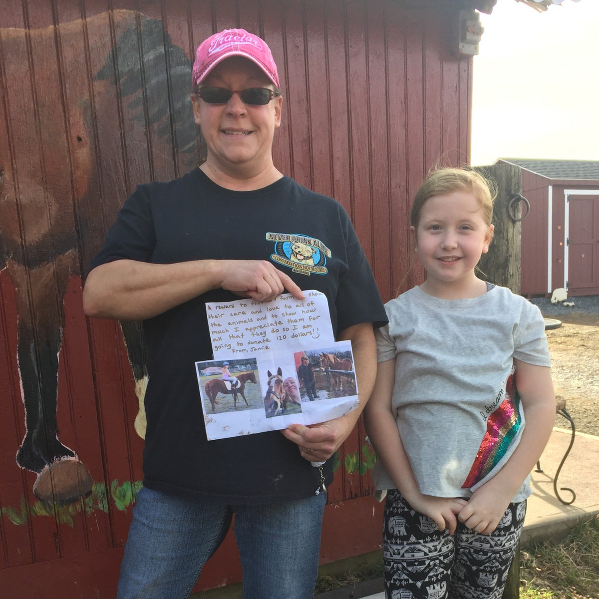 An adult in a farm T-shirt beside a young girl at the barn's cow mural, holding up her handwritten letter pledging a $120 donation