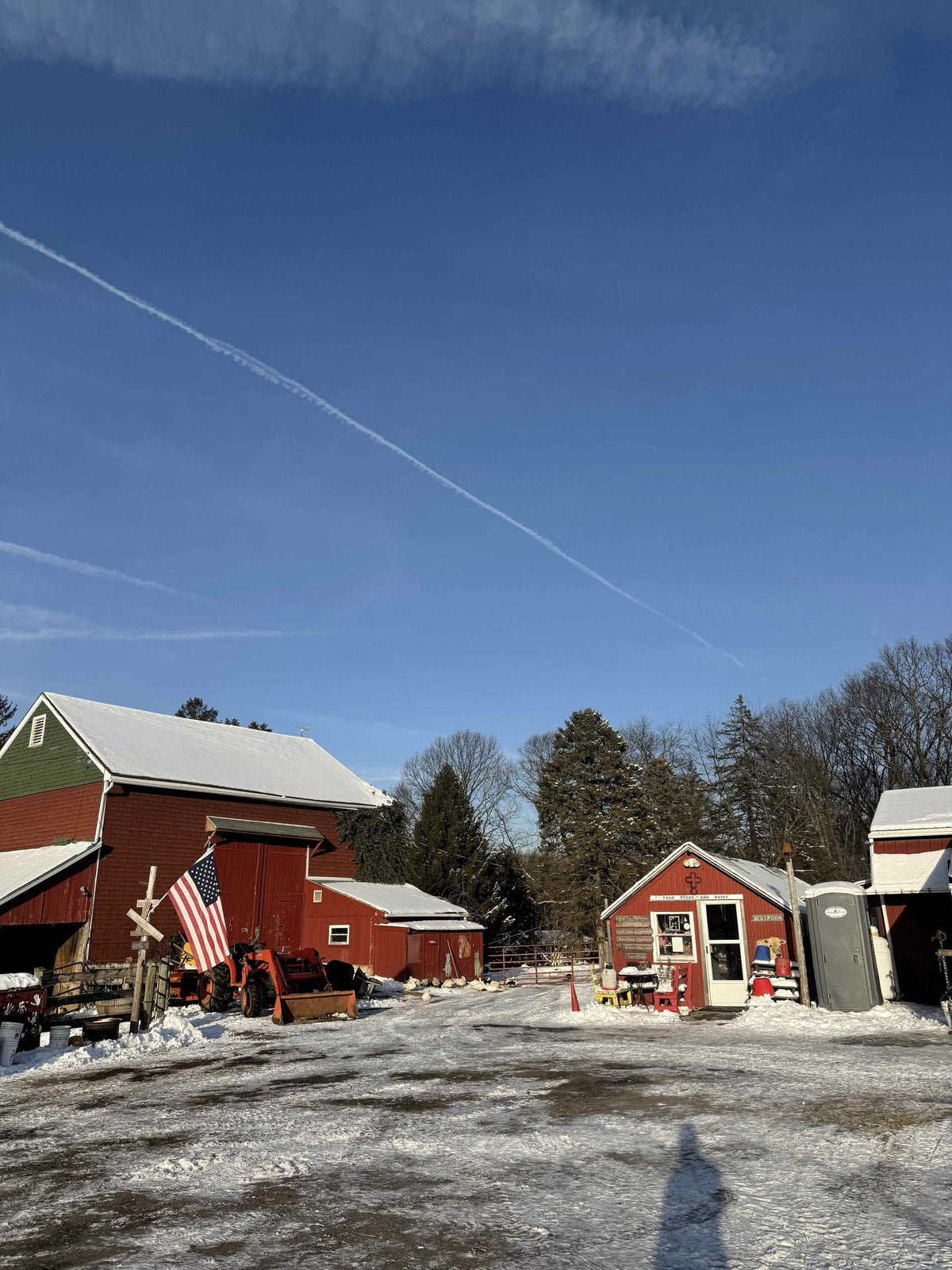 Farmyard after snow: red bank barn with a US flag, an orange Kubota loader tractor, and the small red Farm Store and Dairy under deep blue sky