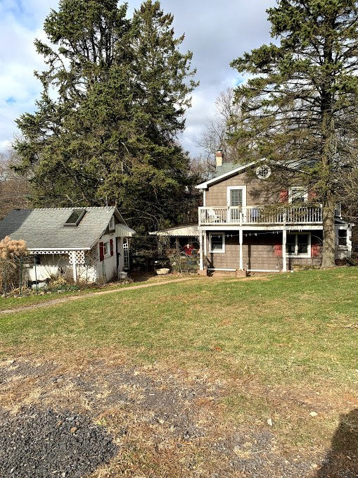 The two-story shingled farmhouse with a covered porch and upper balcony seen across a lawn, framed by tall evergreens, in winter