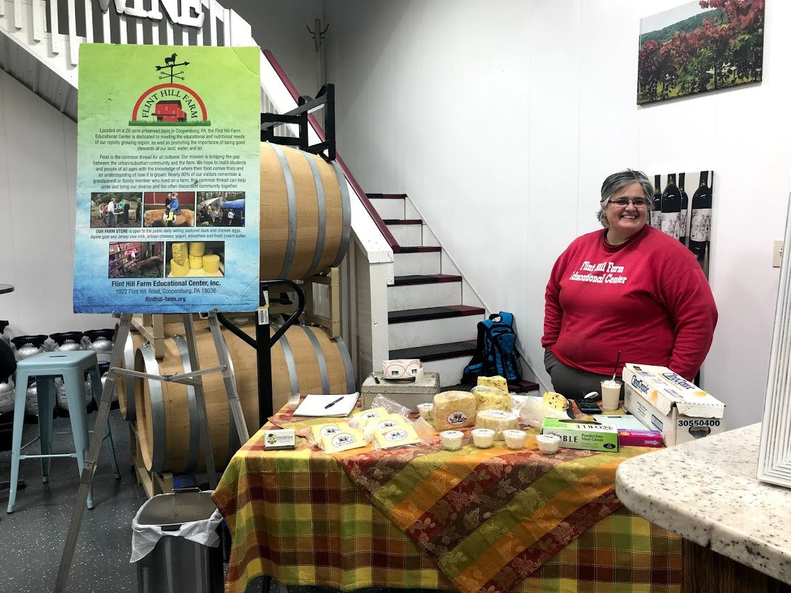 An adult in a Flint Hill Farm Educational Center sweatshirt behind a cheese-tasting table on a plaid cloth, in a room with wine barrels