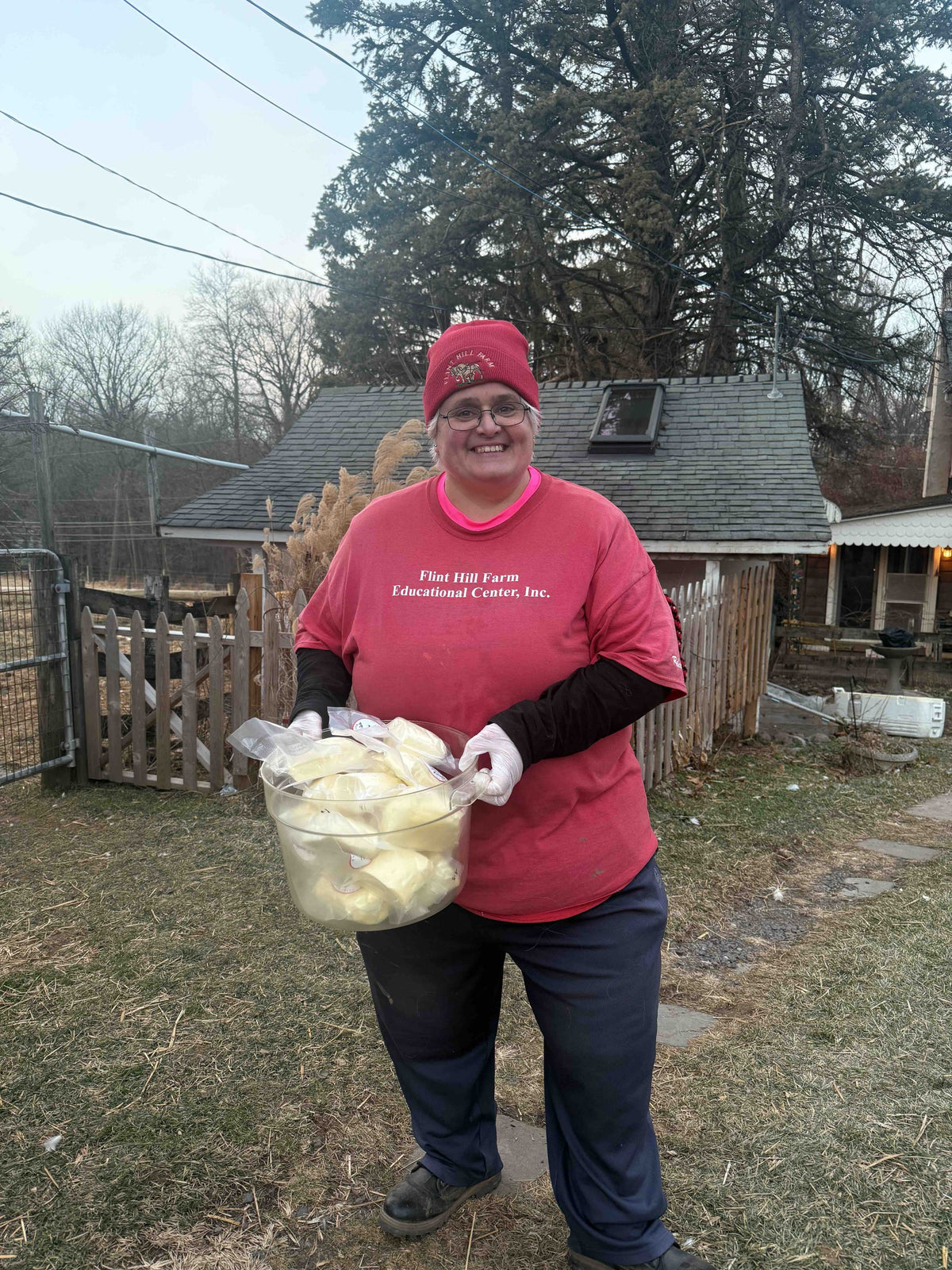 An adult in a Flint Hill Farm Educational Center T-shirt and food gloves carrying a tub of vacuum-bagged fresh white cheese across the yard at dusk