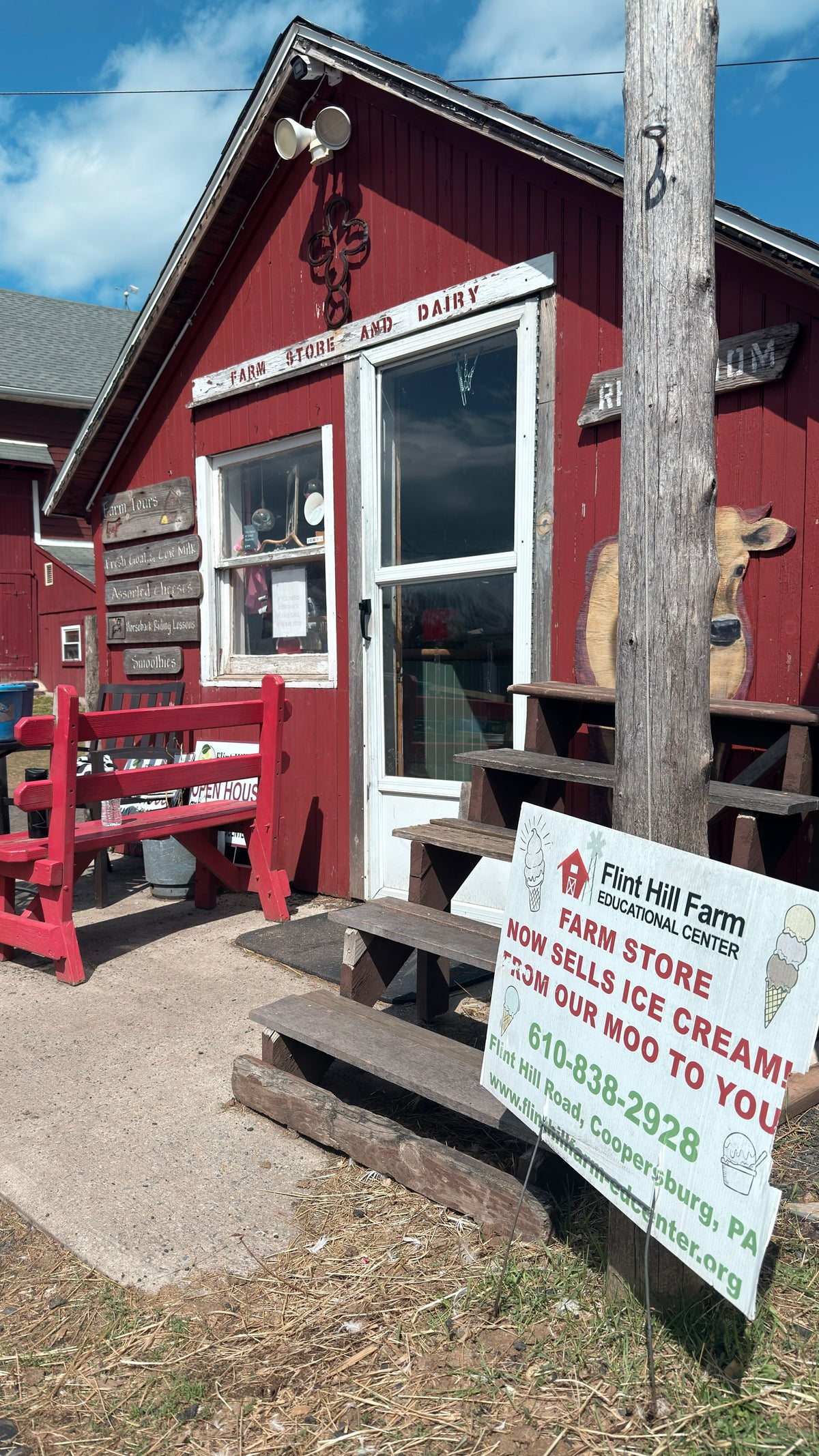 Front of the red board-and-batten Farm Store and Dairy with hand-lettered signs, a painted wooden cow cut-out, red benches and an ice cream sign