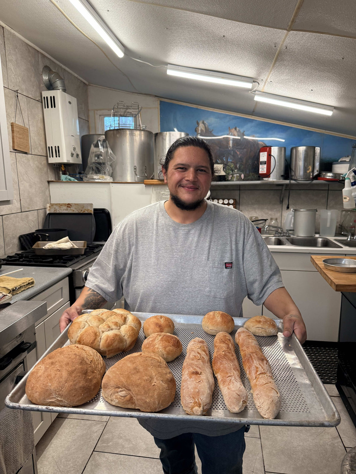 An adult in a gray T-shirt holding a sheet pan of fresh bread, boules, dinner rolls and baguettes, in the farm's tiled kitchen