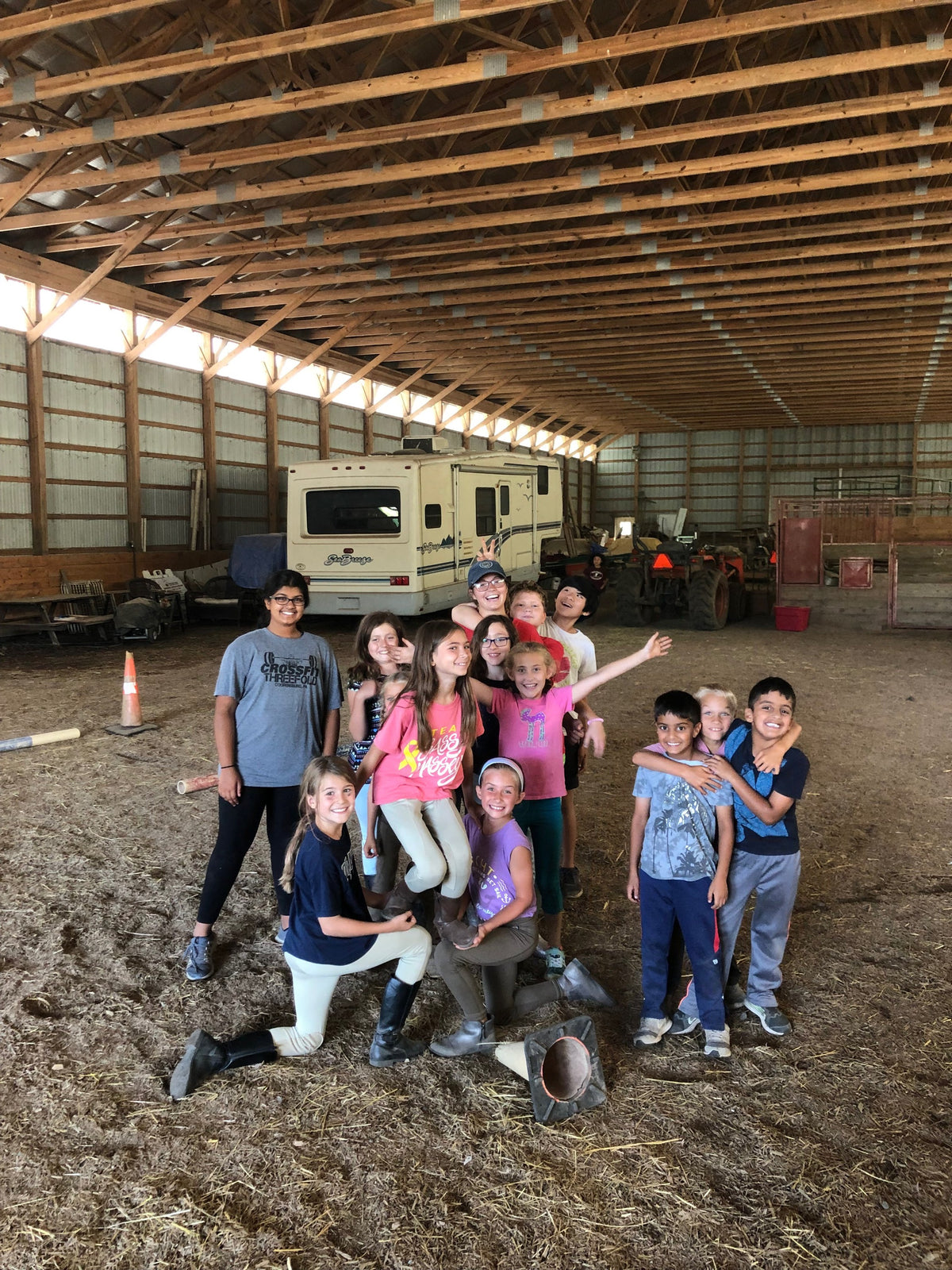 About ten children and teenagers posed in the barn aisle in front of a camper van, several in riding breeches and boots, all smiling