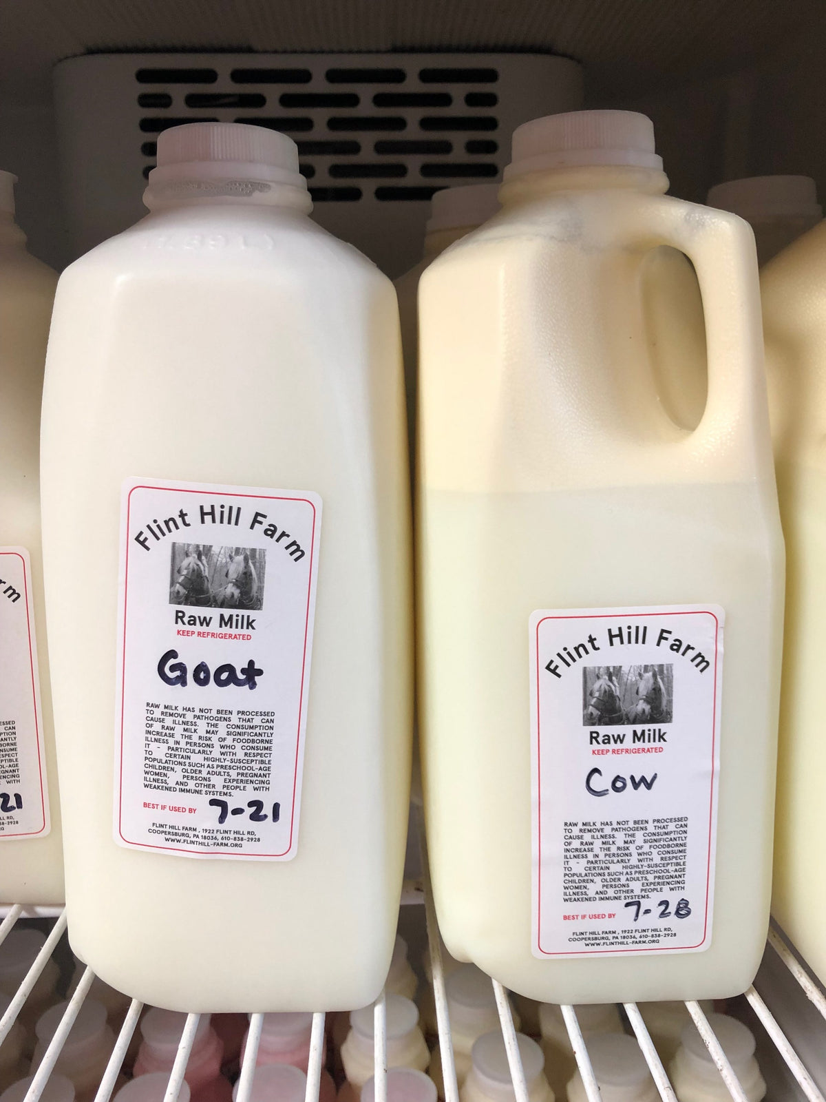 Two half-gallon jugs of Flint Hill Farm raw milk on a store fridge shelf, hand-marked Cow and Goat, with raw-milk warning labels