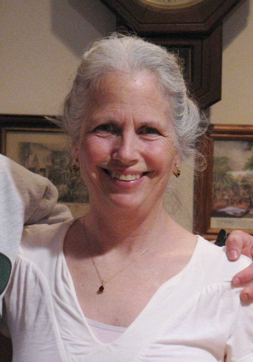 Indoor portrait of an older adult with long gray hair pinned back, smiling in a white top and small pendant, framed prints and a clock behind