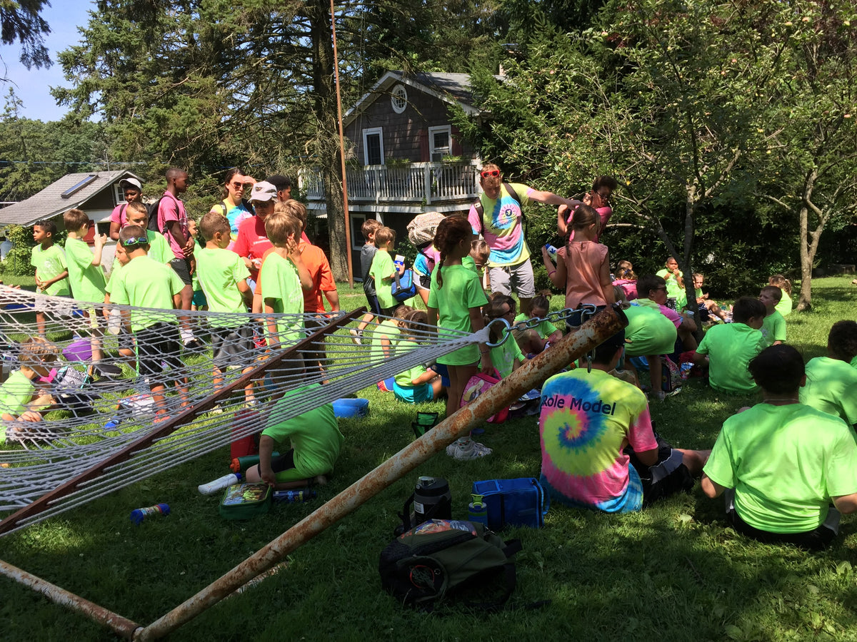 Large group of summer campers in matching lime-green T-shirts on the farmhouse lawn with backpacks and lunch boxes, adult helpers among them