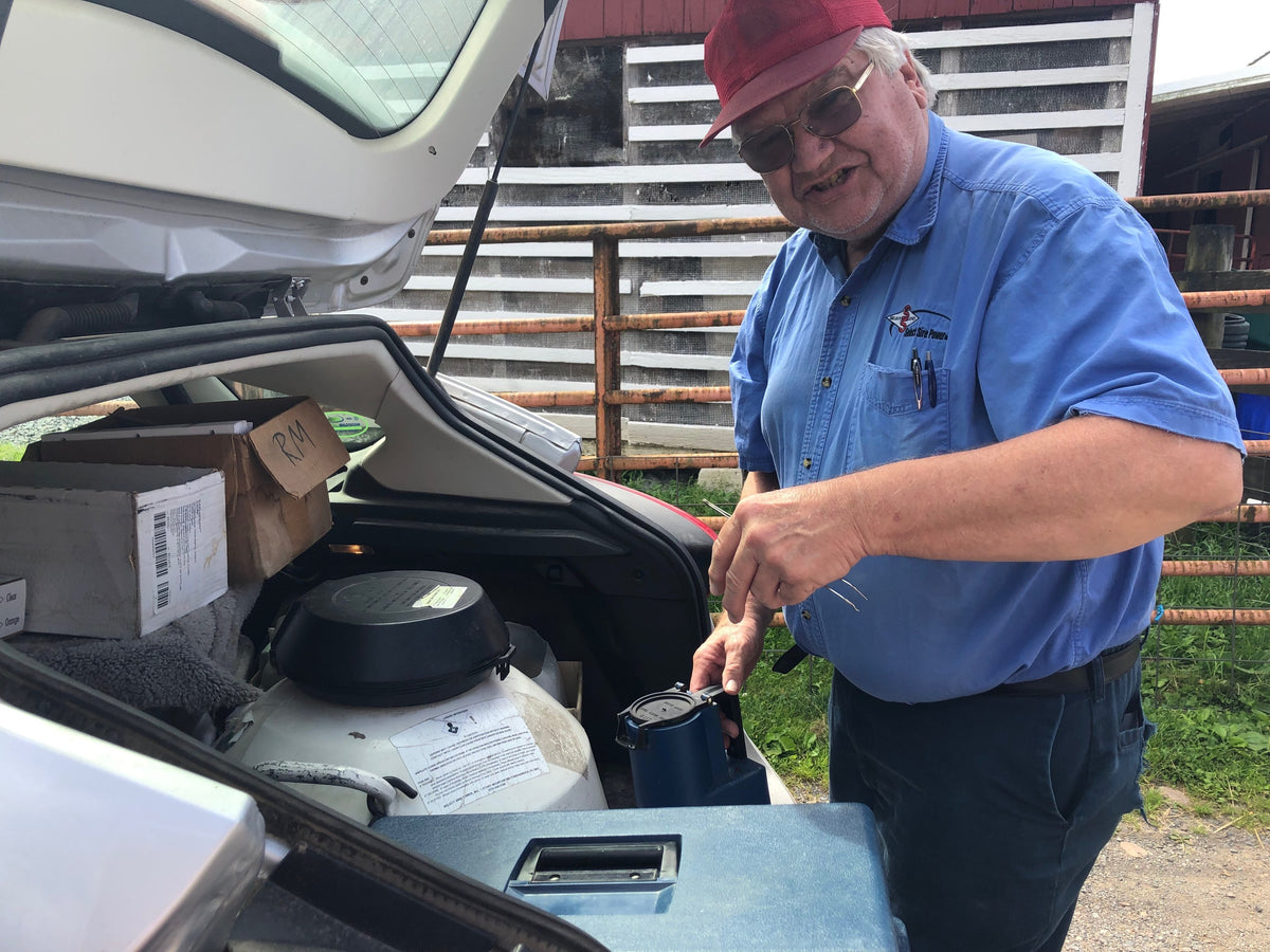 An older adult in a blue work shirt lifting a canister from a liquid-nitrogen breeding tank in a car's open trunk, the barn behind