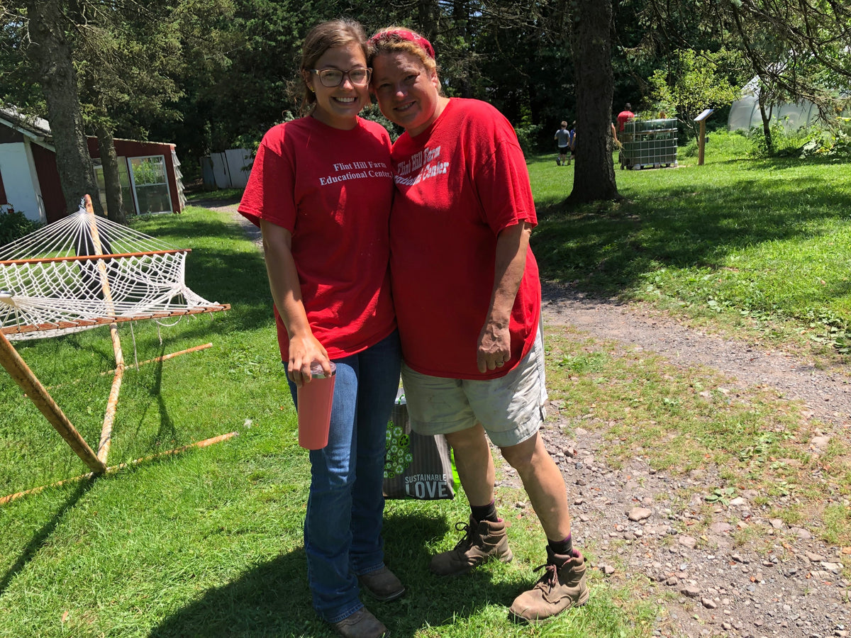 Two people in matching red Flint Hill Farm Educational Center T-shirts cheek-to-cheek on the lawn, smiling, red outbuildings behind