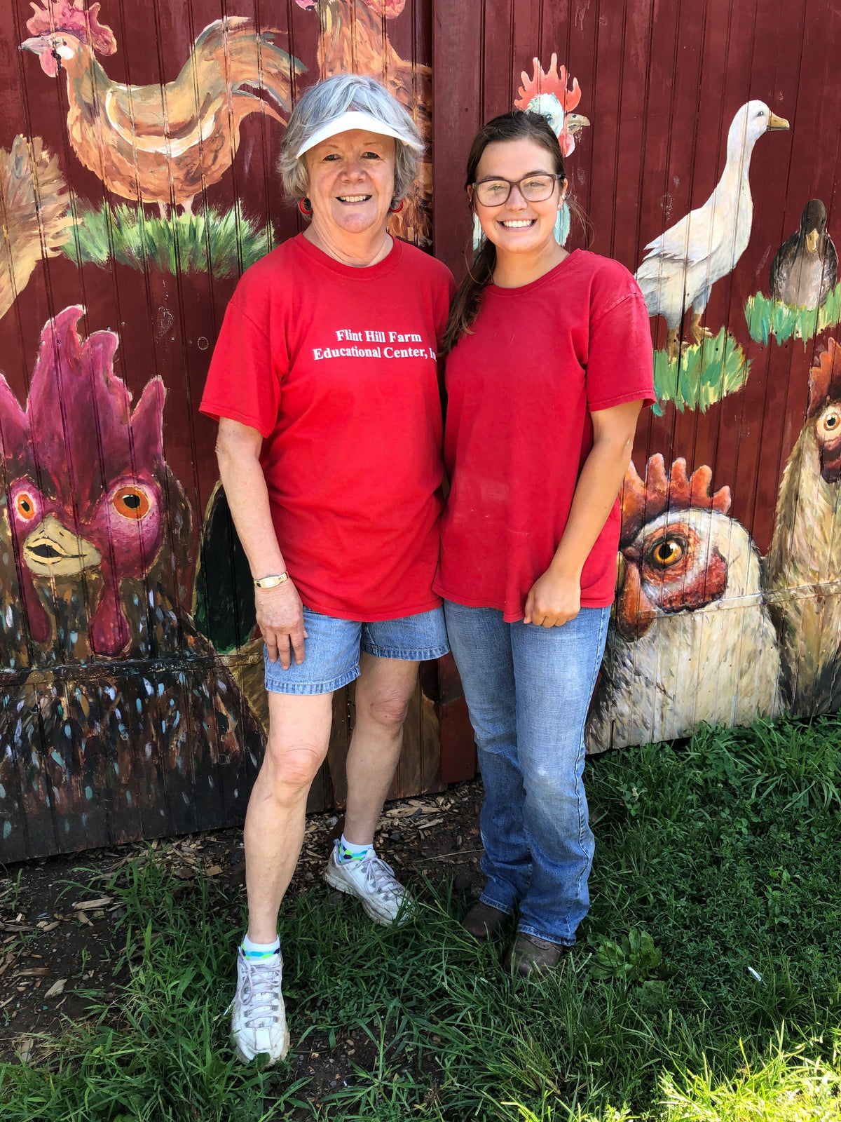Two people in red Flint Hill Farm Educational Center T-shirts smiling as they sit against the barn wall below its life-size chicken and duck mural