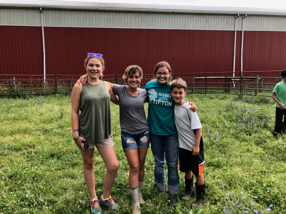 Four young people in muddy boots and work clothes standing arm-in-arm in a flowering grass paddock in front of a long red-and-white barn