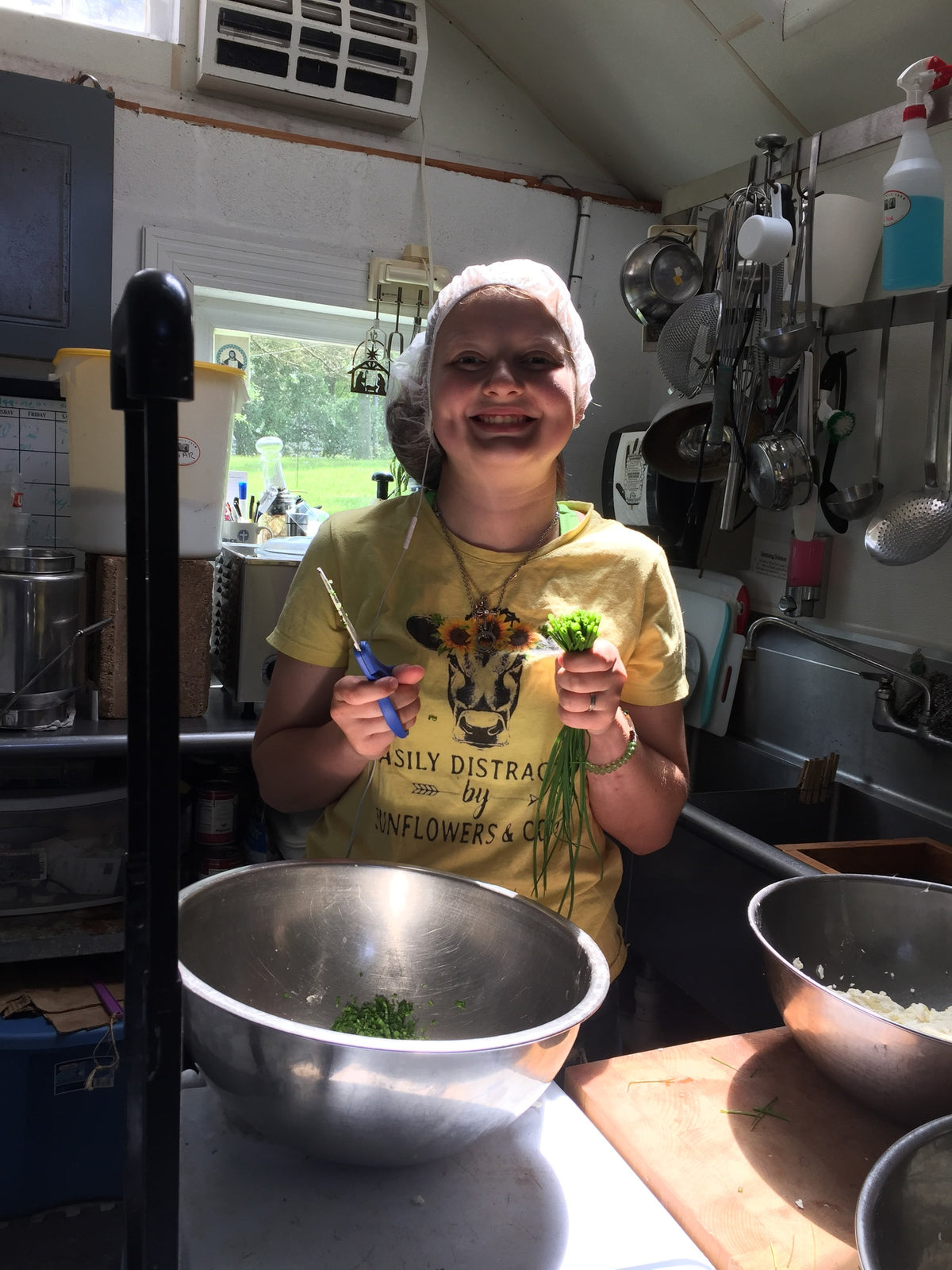 A person in a hairnet in the farm kitchen, grinning while snipping fresh chives into a stainless bowl, a bowl of fresh white curd behind