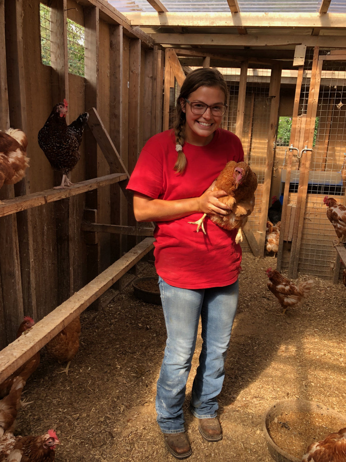 A person in a red Flint Hill Farm Educational Center T-shirt holding a buff hen and smiling in the chicken house, more hens all around