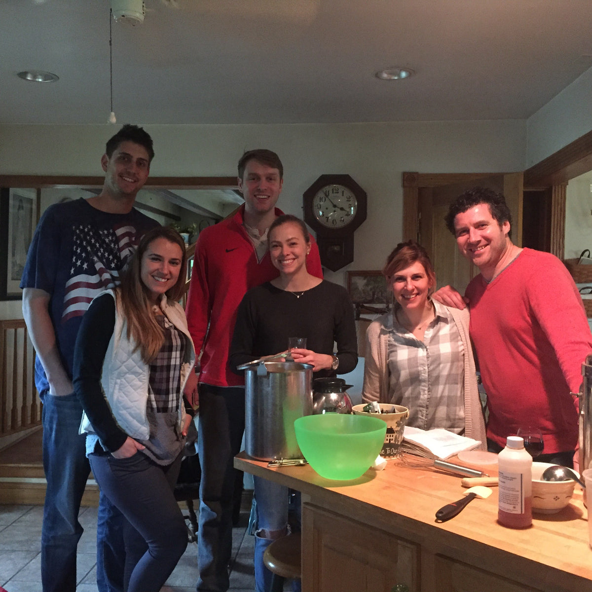 Six adults smiling around a farmhouse kitchen island set up for cheese-making, with a large stockpot, whisks and an open recipe book