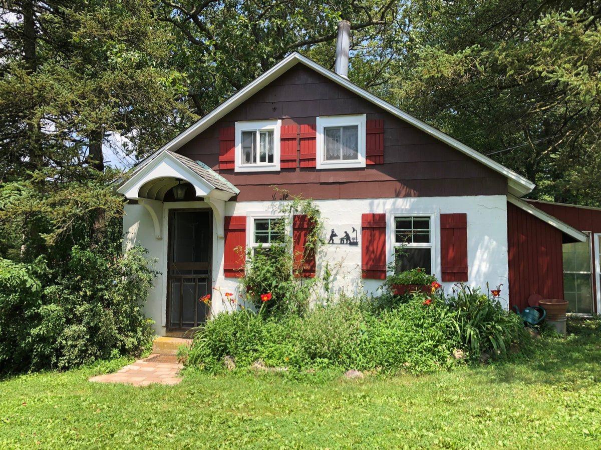 Small farmhouse in summer, dark red shingles over white stucco, red shutters, orange daylilies along the front, a brick path and shade trees