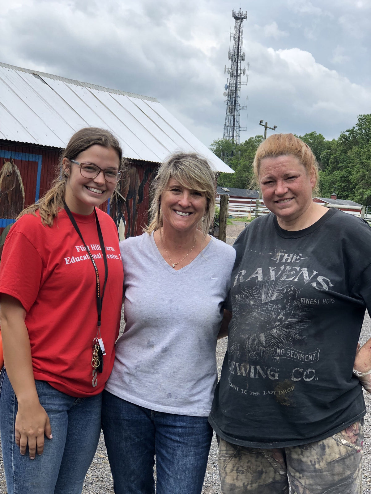 Three adults arm-in-arm on the gravel yard, smiling, one in a red Flint Hill Farm Educational Center T-shirt, the red barn's horse mural behind