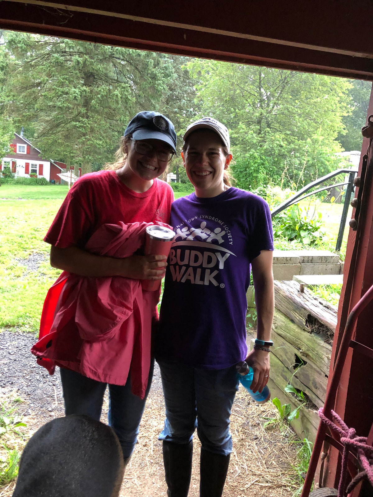 Two adults smiling in a barn doorway, each holding a drink bottle, one in a purple charity walk T-shirt, a red farm building behind