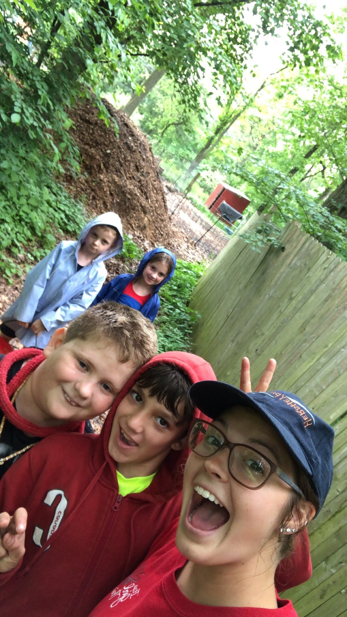 Tilted selfie of an adult in a cap and glasses with three children in rain jackets on a wooded trail beside a tall wooden fence