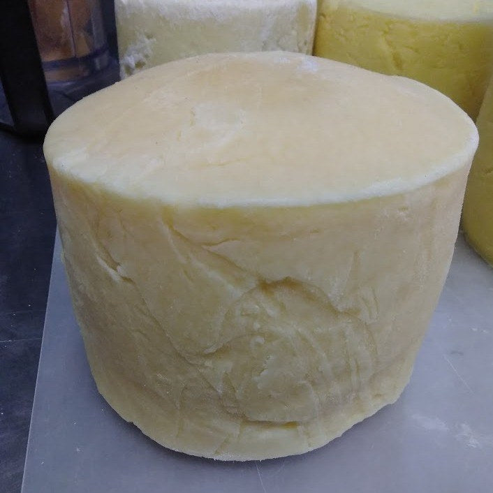 A whole wheel of pale-yellow parmesan-style hard cheese on a stainless steel work surface, two more wheels behind it