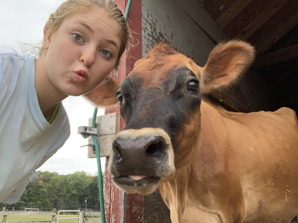 A young woman cheek-to-cheek with a fawn Jersey cow in a barn doorway, the cow looking straight into the lens with its tongue out