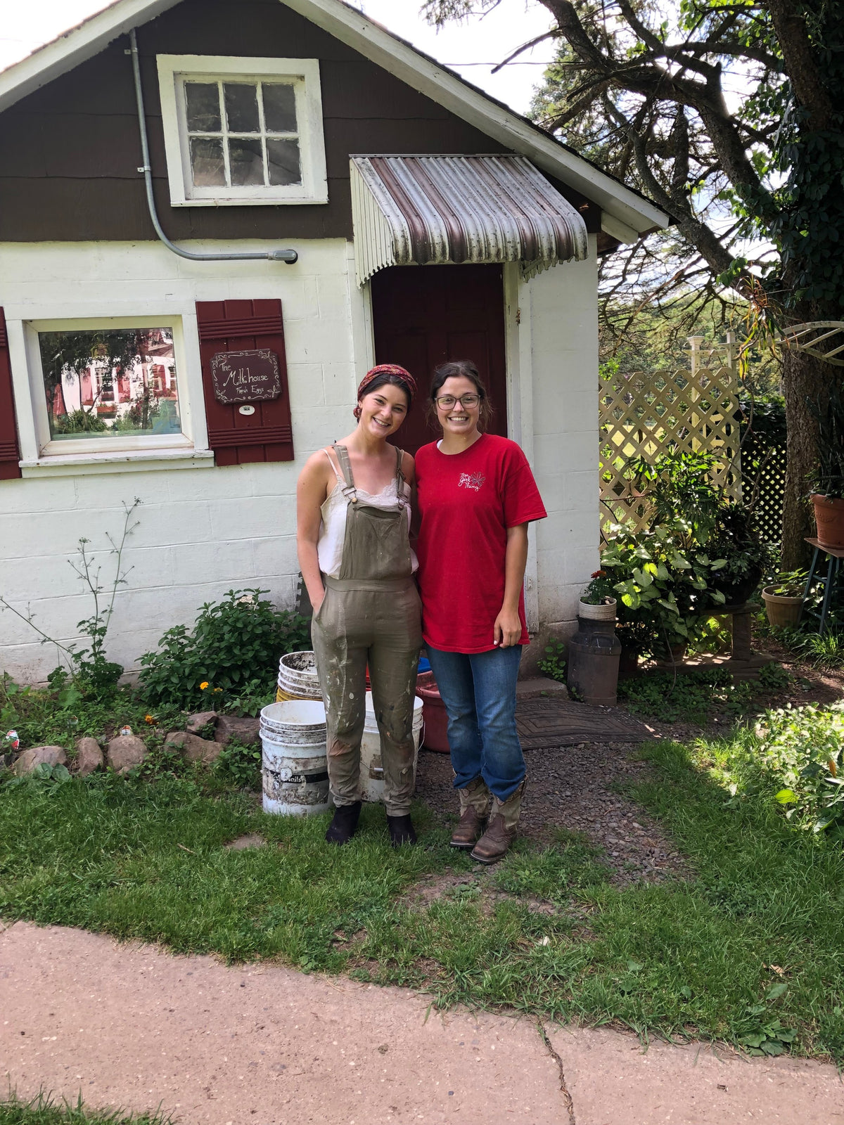 Two adults in work clothes side by side outside the small white Milkhouse building, under its hand-lettered The Milkhouse, Fresh Eggs sign