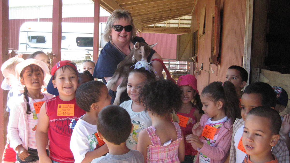 A dozen preschool-age children crowded under a barn overhang around an adult holding a young brown-and-white goat